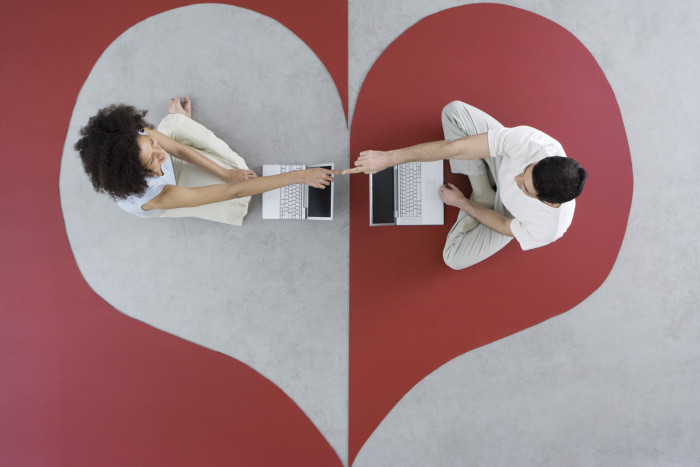 Couple sitting face to face with laptop computers on heart shape, touching fingers, overhead view